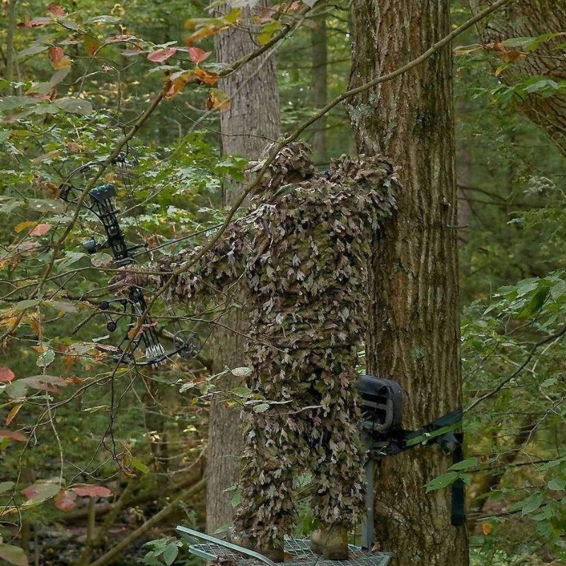 A wilderness researcher blending seamlessly into a forest environment next to a tree trunk using the wind-resistant Solid-Shell 3D Leafy Suit.