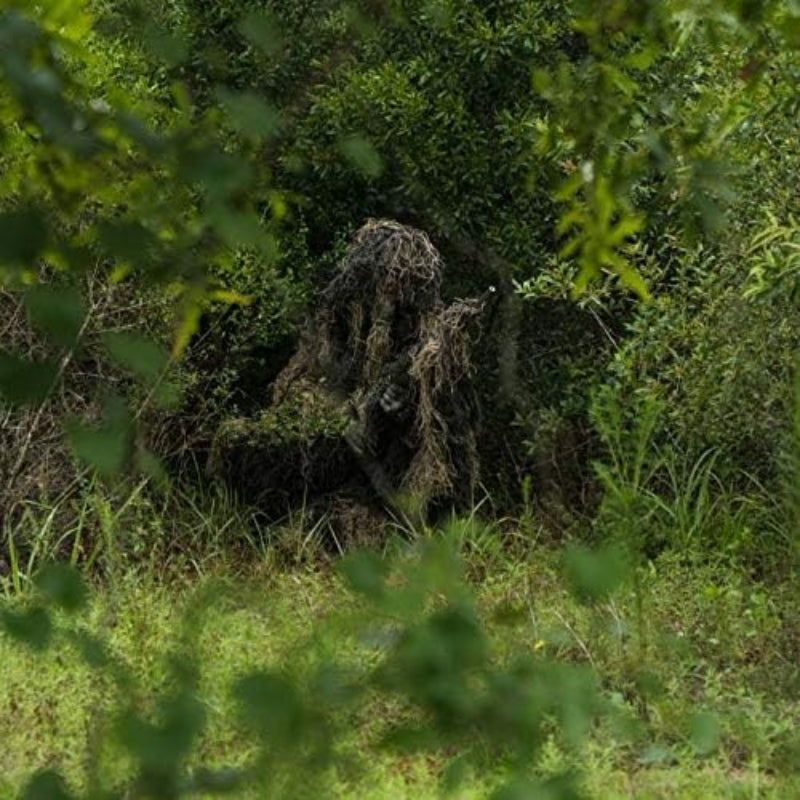 A wildlife photographer crouching in thick green brush, achieving complete profile disruption with the zero-shed Pro-Series synthetic thread ghillie suit.