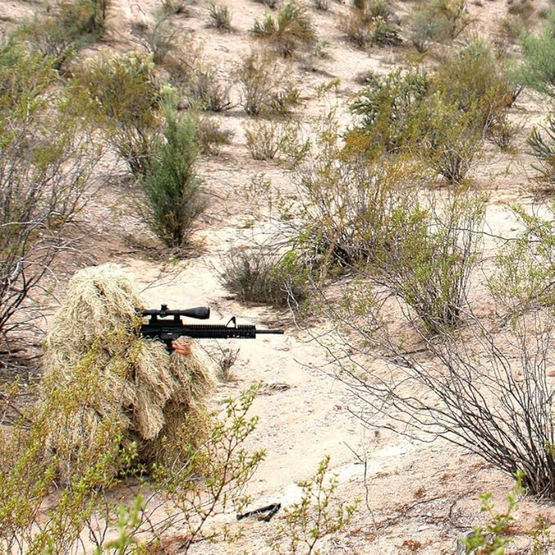 A wilderness researcher wearing the 100% weather-resistant Ghost Ghillie suit, adjusting long-focus optics while remaining fully camouflaged in a dry, gravelly environment.