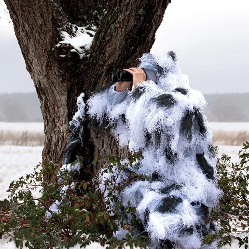 A professional alpine researcher wearing a wind-resistant snow ghillie suit, standing next to a dark tree trunk to conduct cold-weather birdwatching.