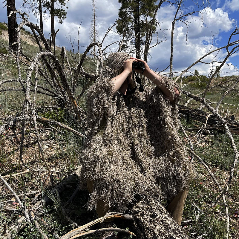 A tracker wearing the Dry Grass Ghost Ghillie Poncho while using binoculars in a dry, post-burn forest environment.