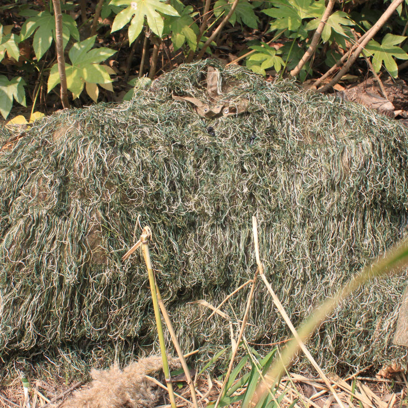 Extremely low-profile prone concealment in a green forest hollow using the Woodland variant Ghost Ghillie Poncho