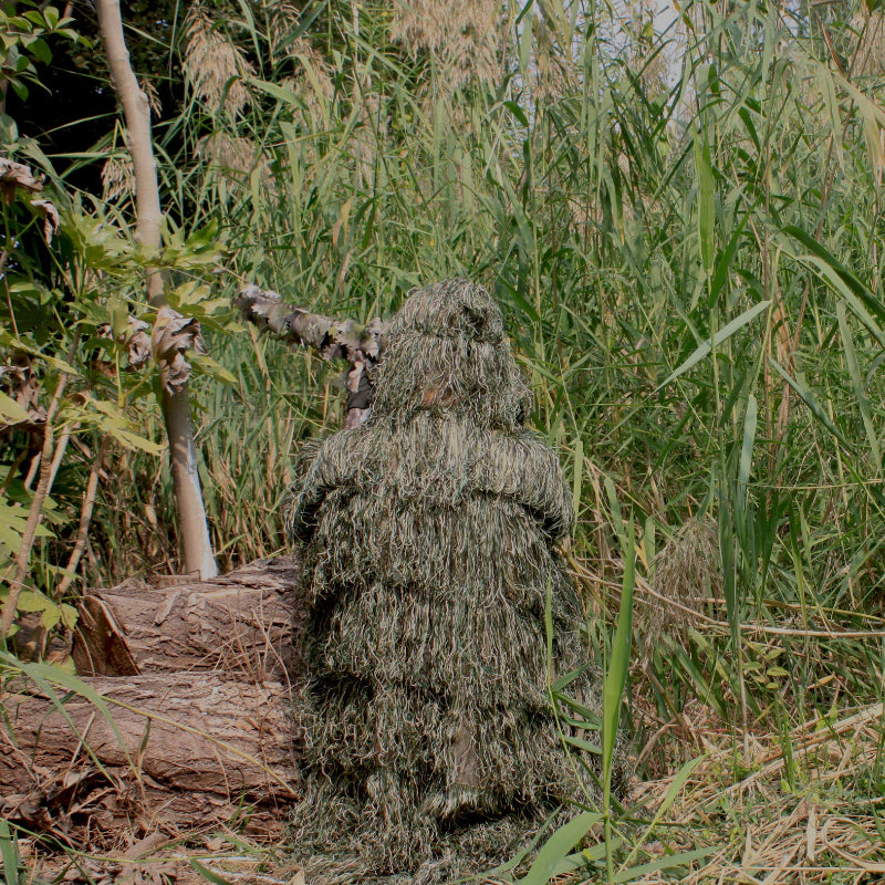 Seated back view of a tracker wearing the Woodland variant Ghillie Poncho, blending into tall lakeside reeds and green brush