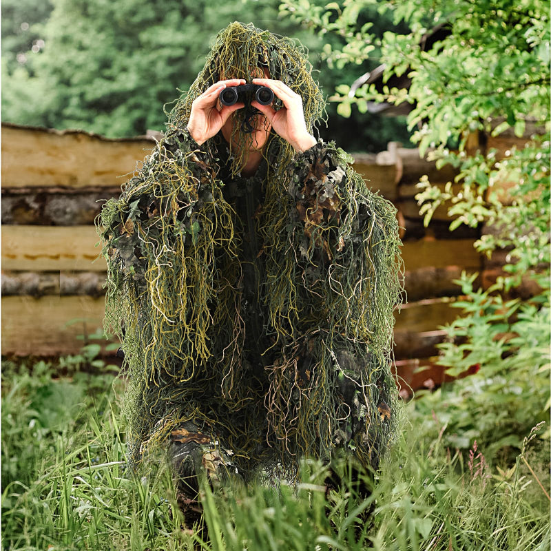 A tactical tracker using binoculars while fully concealed in dense greenery wearing the Ghost Hybrid Ghillie Suit.